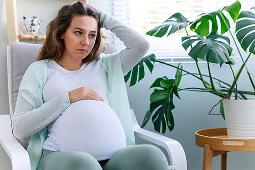 Pregnant woman in her living room, holding both her head and belly
